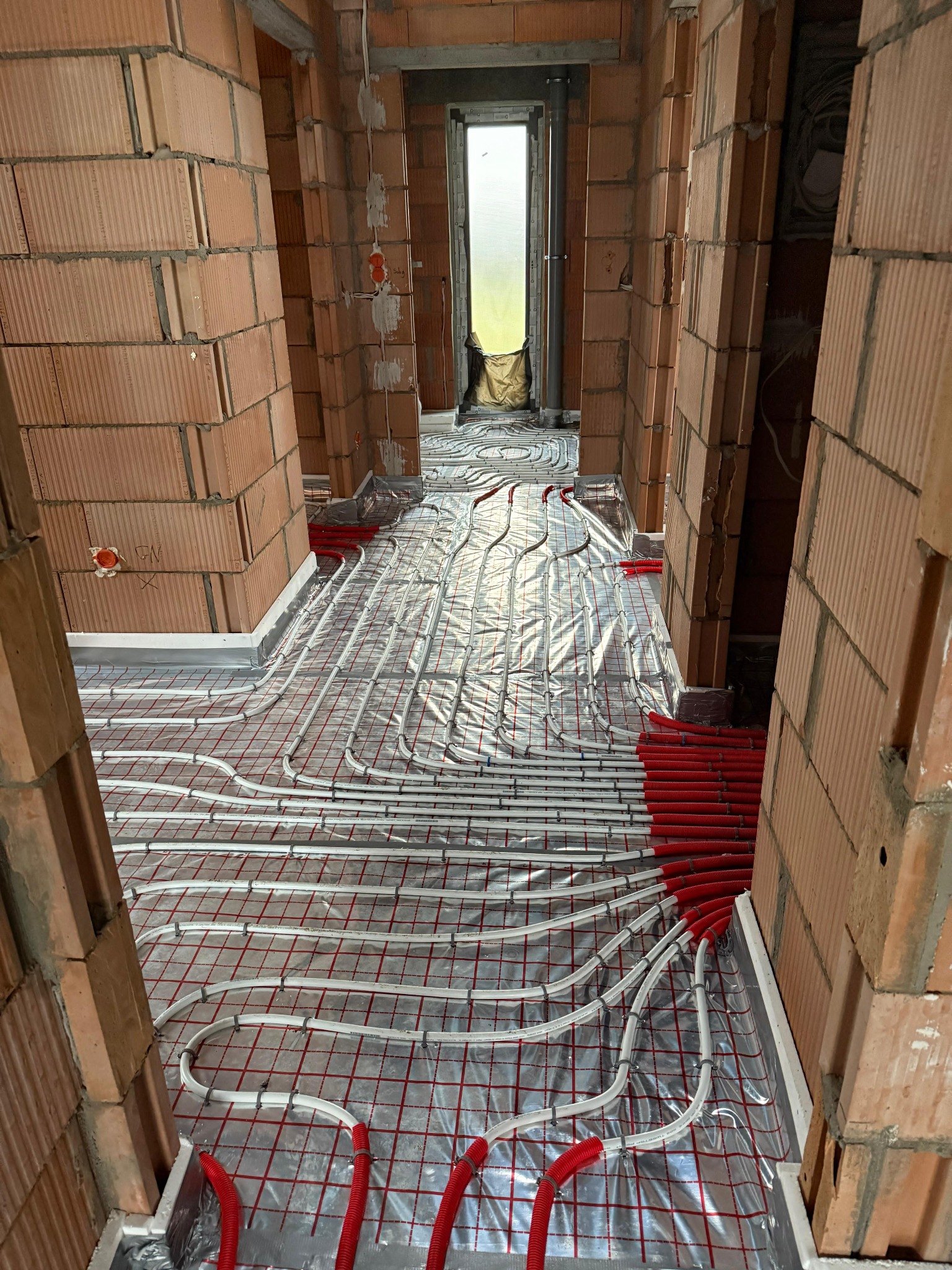 Instalacja ogrzewania podłogowego w surowym wnętrzu z czerwonej cegły. Białe rurki grzewcze ułożone na srebrnej folii z czerwoną siatką, widok w perspektywie korytarza.