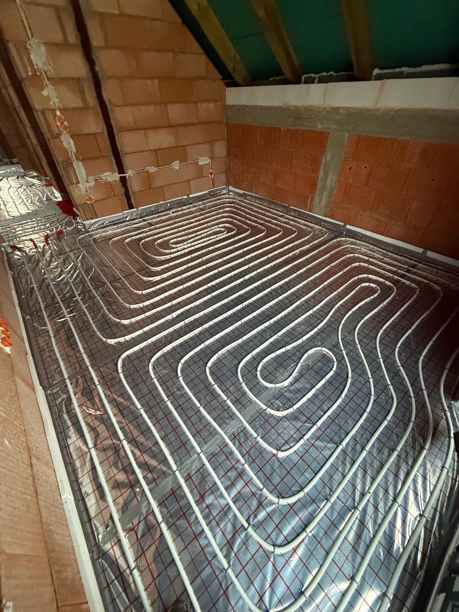 Ułożone rury ogrzewania podłogowego na folii izolacyjnej w pomieszczeniu ze ścianami z cegły. Widok z góry, instalacja w trakcie realizacji.