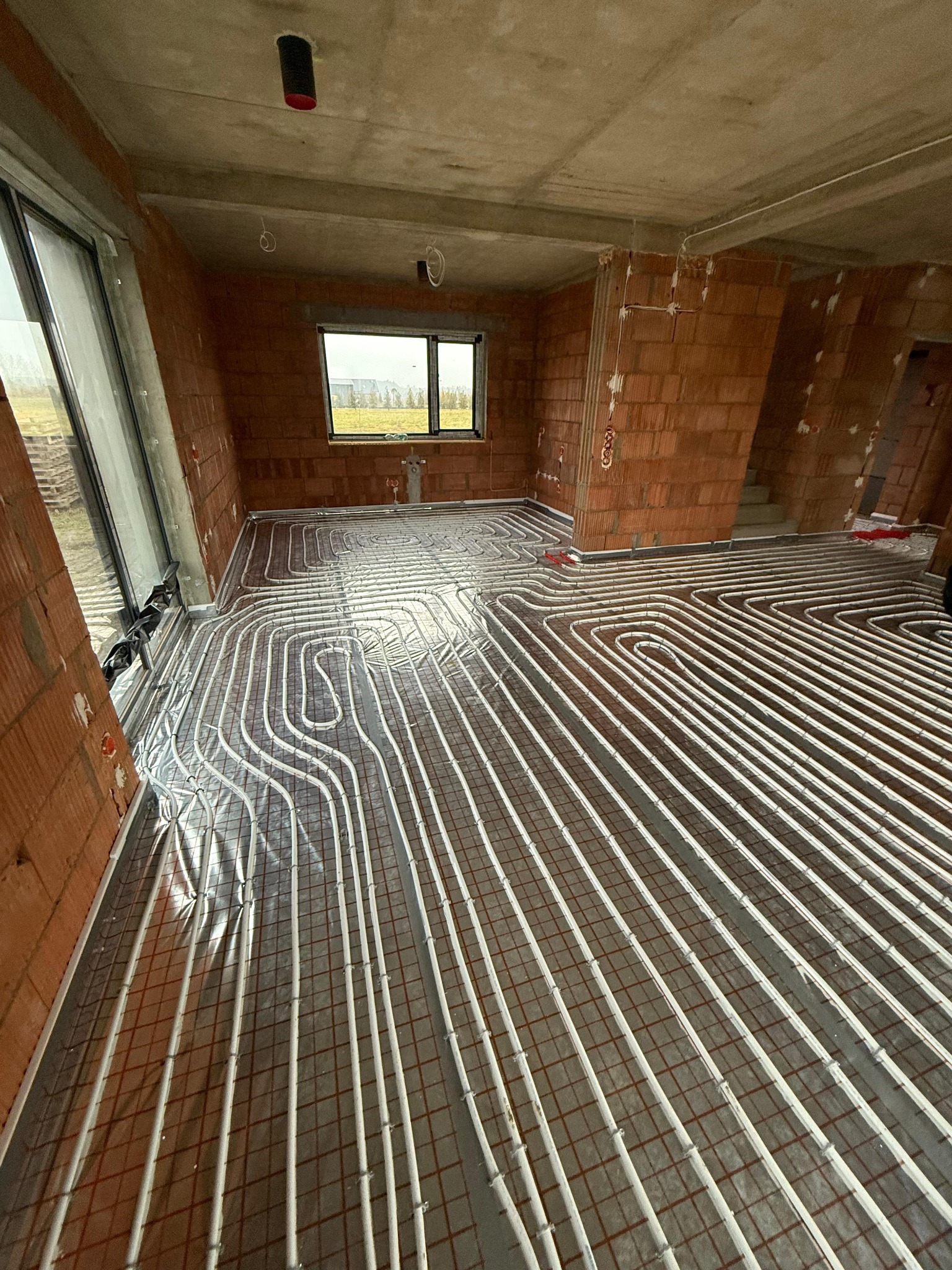 Białe rury ogrzewania podłogowego ułożone na siatce w surowym wnętrzu budynku z ceglanymi ścianami i oknem. Widok z góry, perspektywa zbieżna.