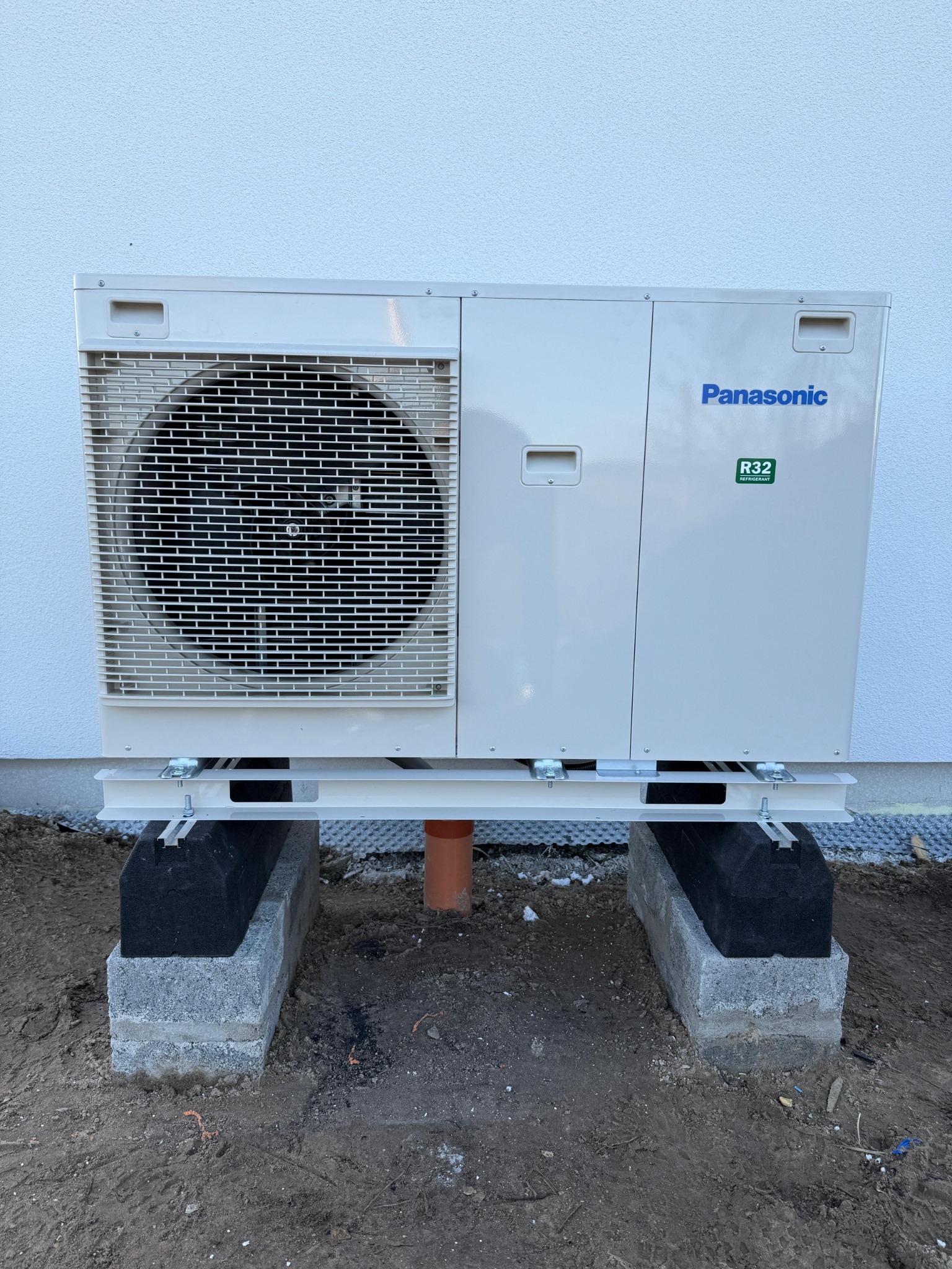Zewnętrzna jednostka pompy ciepła Panasonic zamontowana na betonowych blokach z izolacją, na tle białej ściany budynku. Widoczny wentylator i naklejka R32.