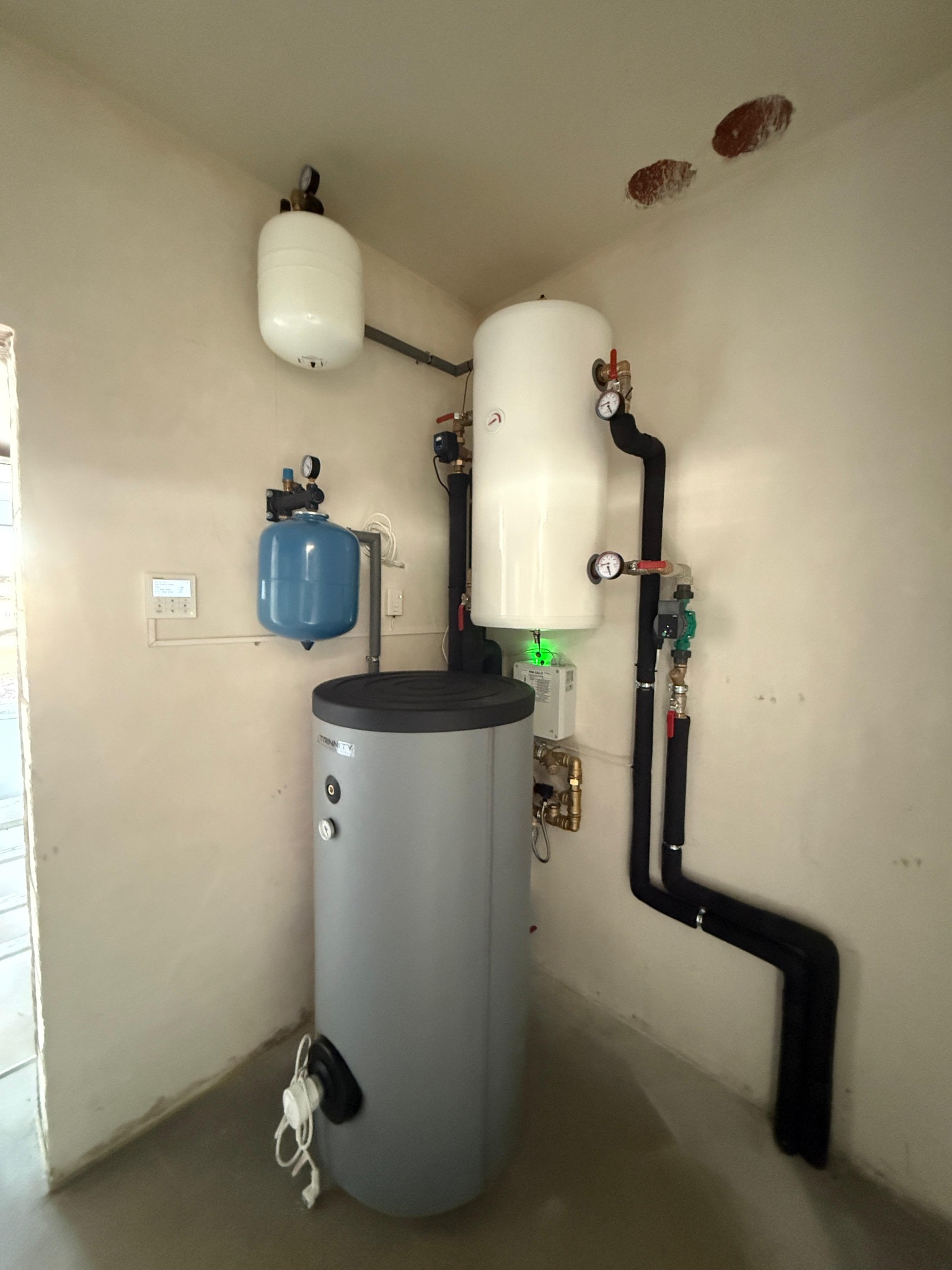 Zestaw instalacji grzewczej w rogu pomieszczenia: szary zbiornik, białe zbiorniki wyrównawcze, czarne rury. Widoczne manometry i zawory. Ściany w odcieniach beżu.