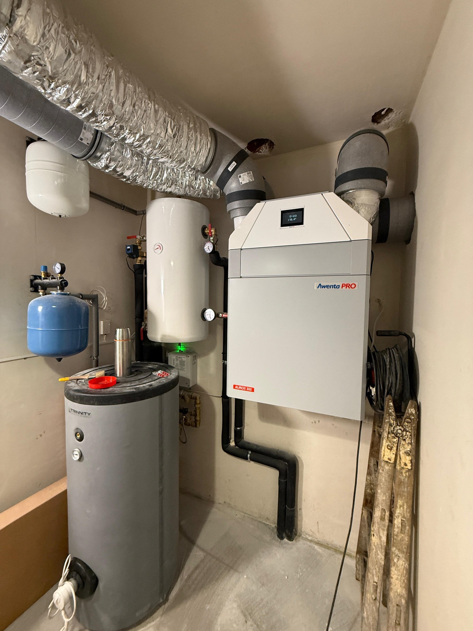 Zestaw urządzeń w kotłowni: rekuperator Aventa PRO, zbiornik buforowy Trinity, naczynia przeponowe i instalacja wentylacyjna z rurami spiro. Widoczne drabiny malarskie.