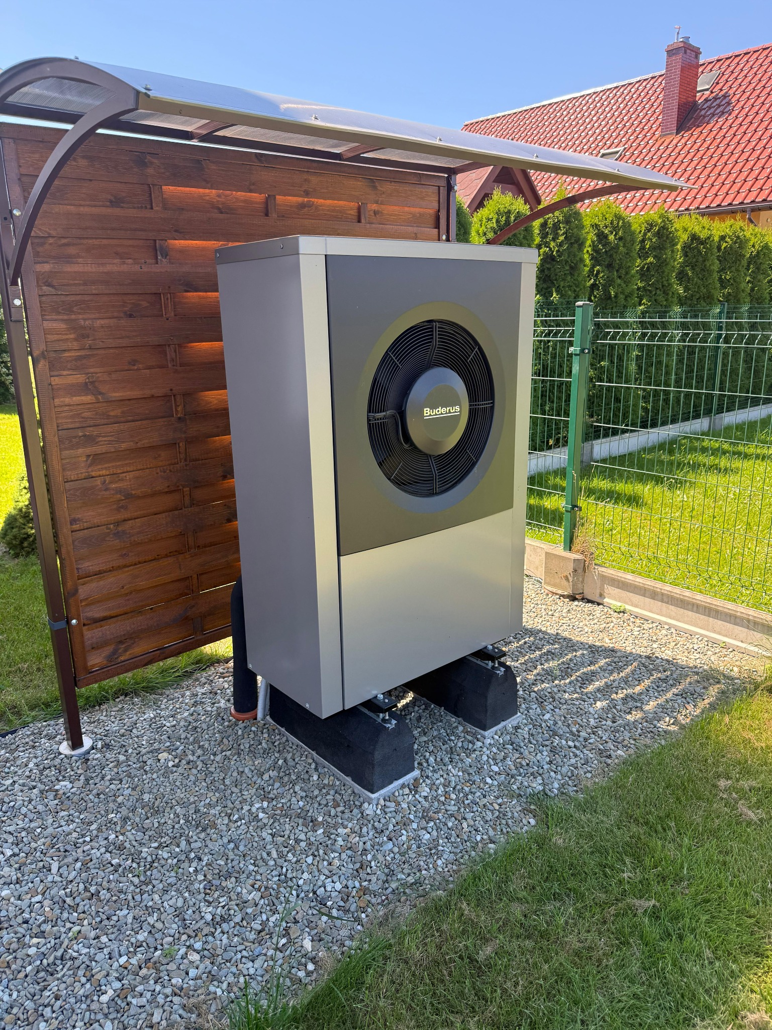 Szara pompa ciepła Buderus na żwirowym podłożu, osłonięta drewnianą konstrukcją z przezroczystym zadaszeniem, na tle zielonego trawnika i ogrodzenia.