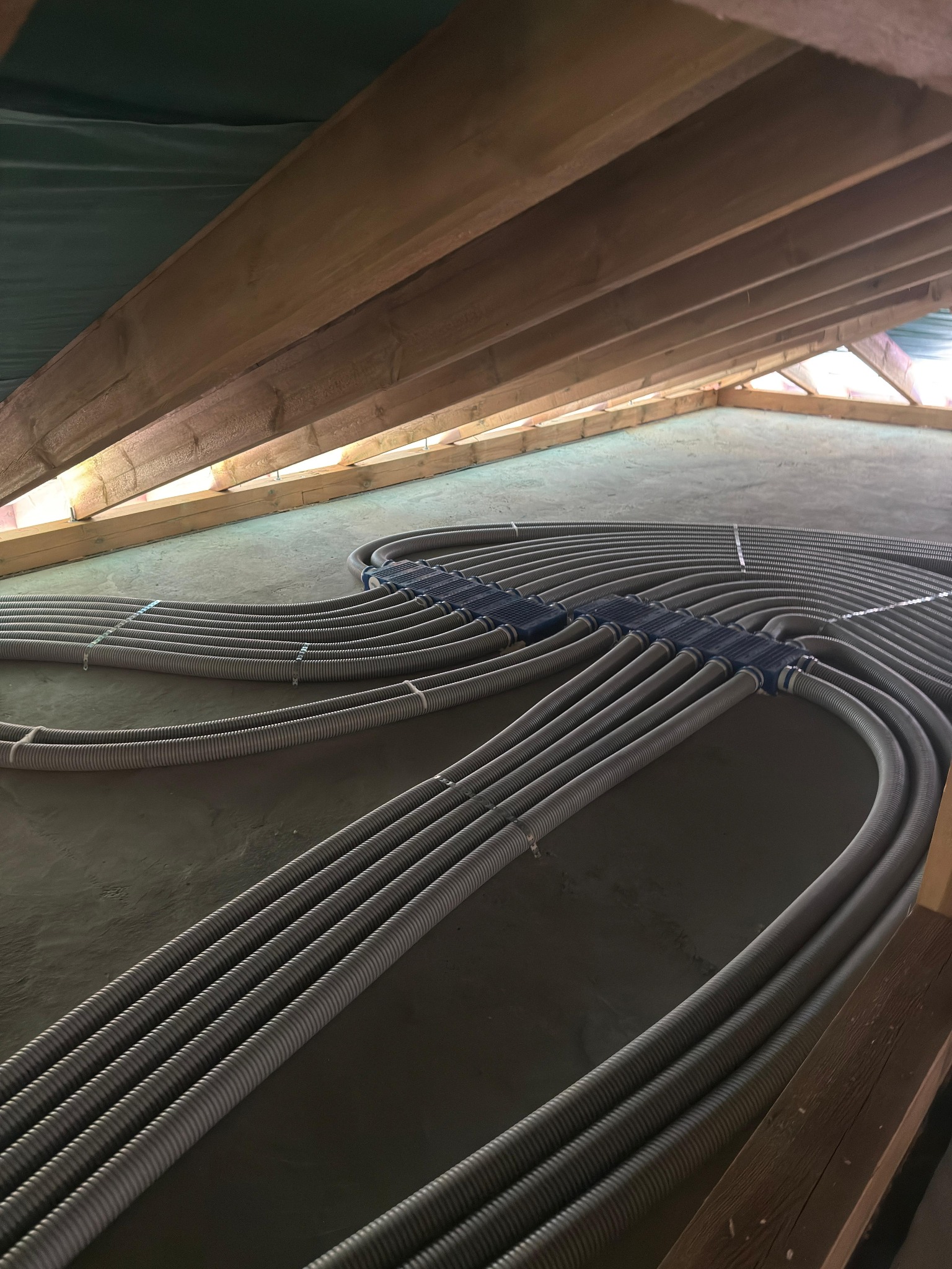 Szare rury instalacji ogrzewania podłogowego, ułożone równolegle na szarej powierzchni, z drewnianą konstrukcją dachu w tle. Widok z góry, szeroki kąt.