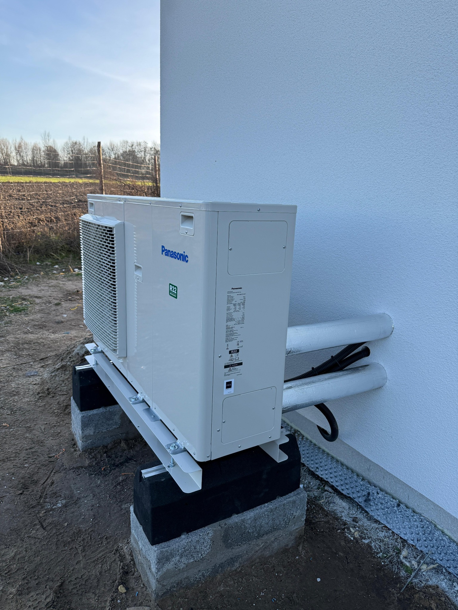 Biała pompa ciepła Panasonic zamontowana na betonowych blokach przy ścianie budynku. Rury w izolacji termicznej wchodzą do budynku. Widok z boku, w tle pole.