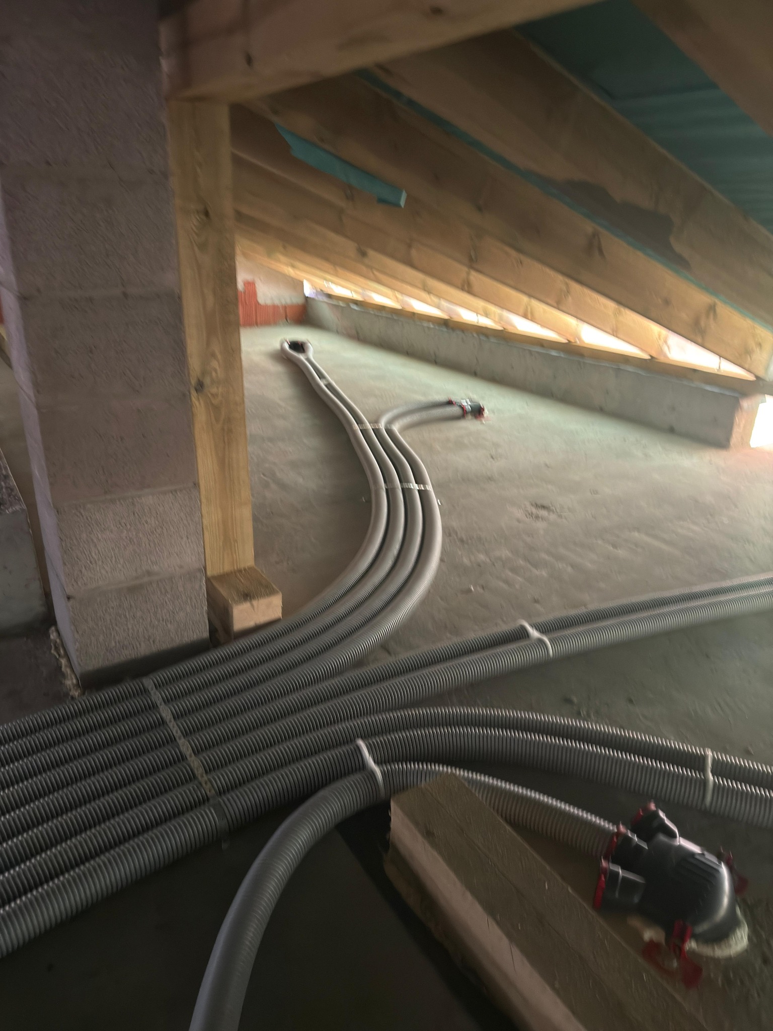 Szare, karbowane rury wentylacyjne ułożone na poddaszu, rozchodzące się w różnych kierunkach. Widoczne drewniane elementy konstrukcyjne dachu i fragment ściany z pustaków.