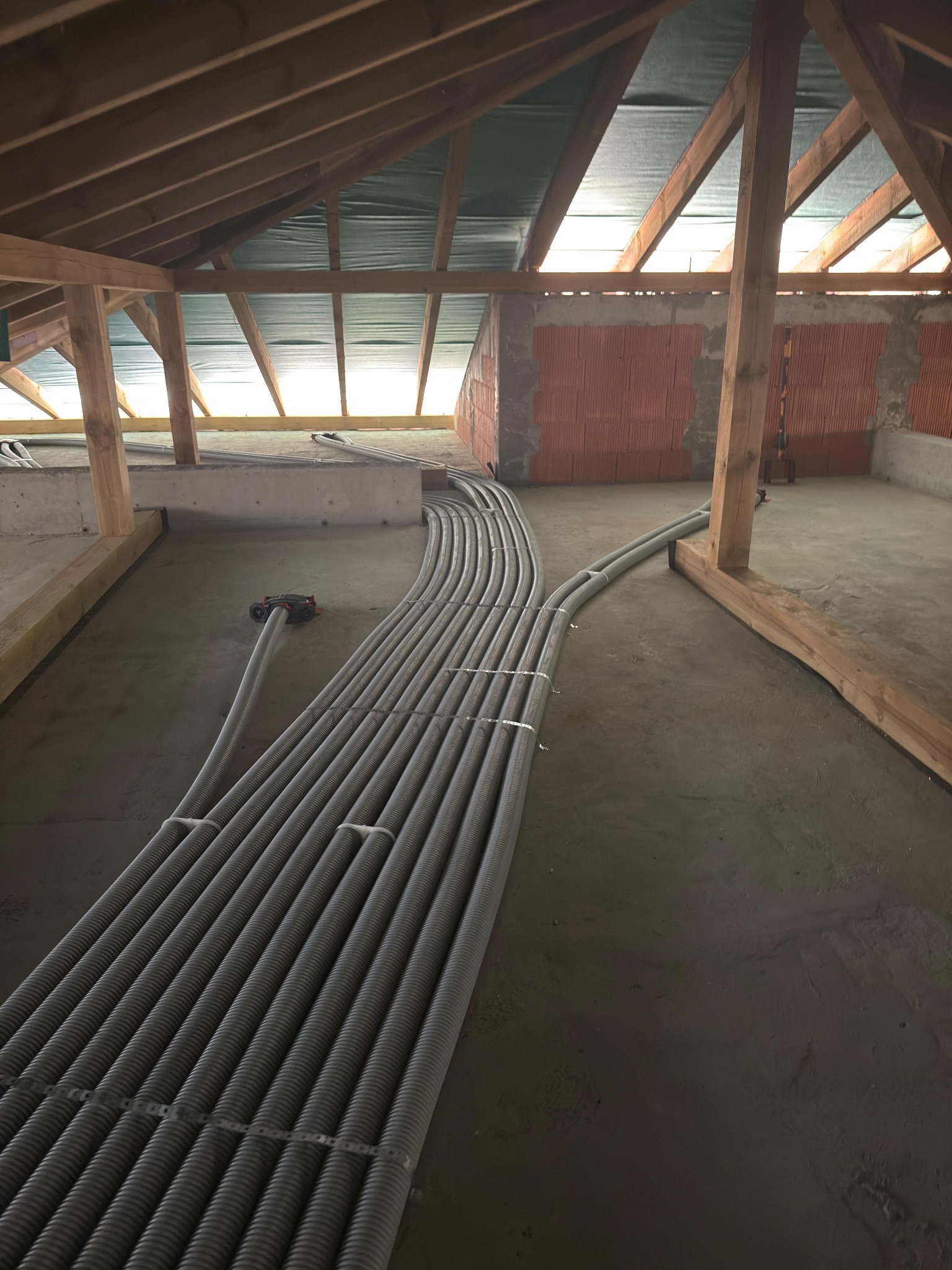 Szare rury instalacyjne ułożone równolegle na podłodze w surowym stanie, widoczne drewniane elementy konstrukcji dachu i ściany z czerwonej cegły.