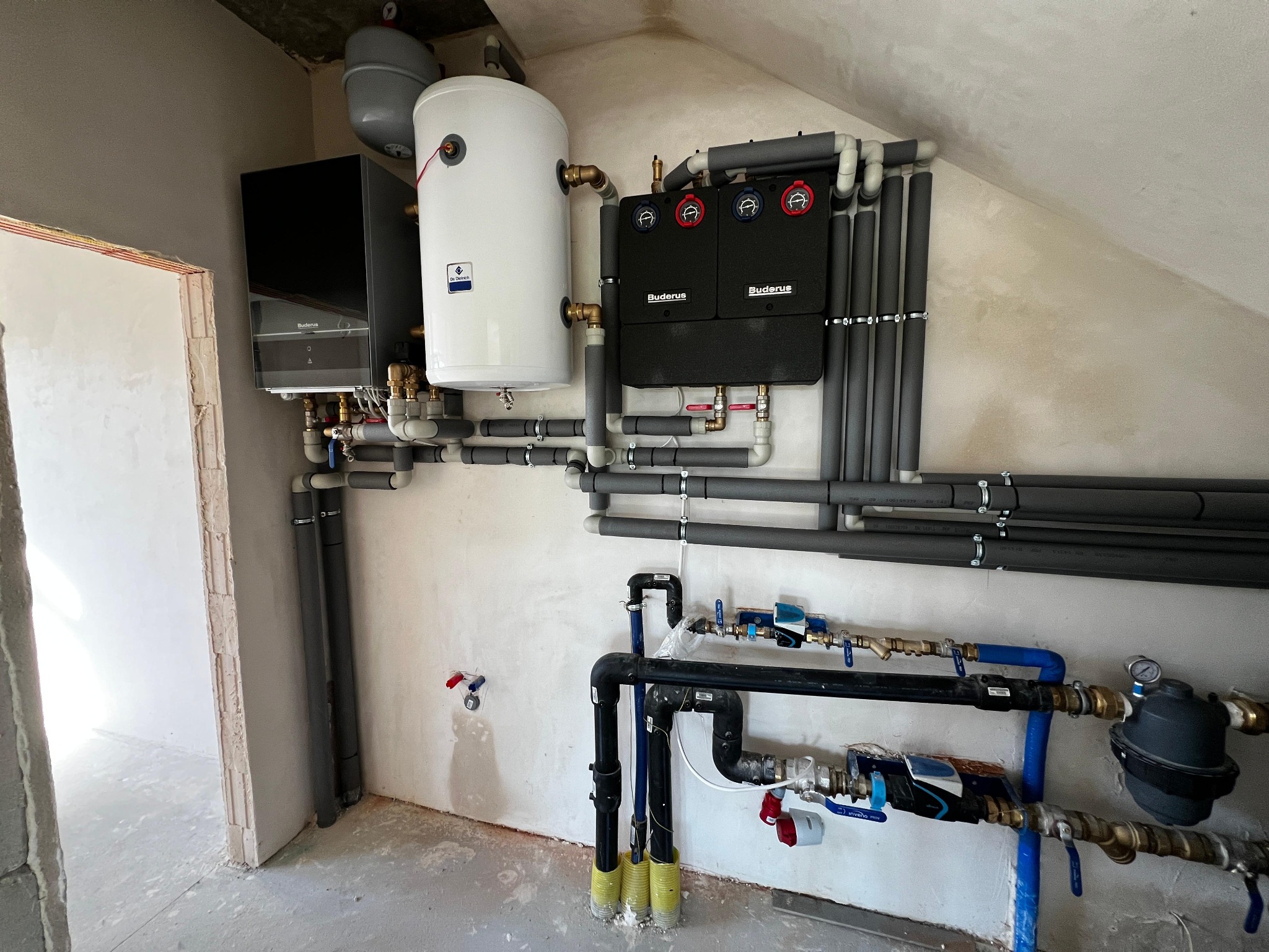 Złożona instalacja grzewcza Buderus w surowym wnętrzu: piec, zasobnik, rozdzielacze i rury z izolacją. Widoczne liczne zawory, manometry i elementy przyłączeniowe.