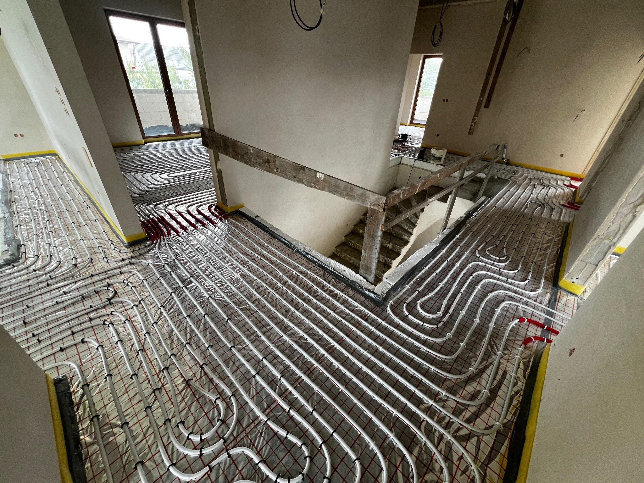 Rozłożona instalacja ogrzewania podłogowego w surowym wnętrzu domu, widoczne białe rury na folii z czerwoną siatką, żółta taśma przy ścianach, schody w tle.
