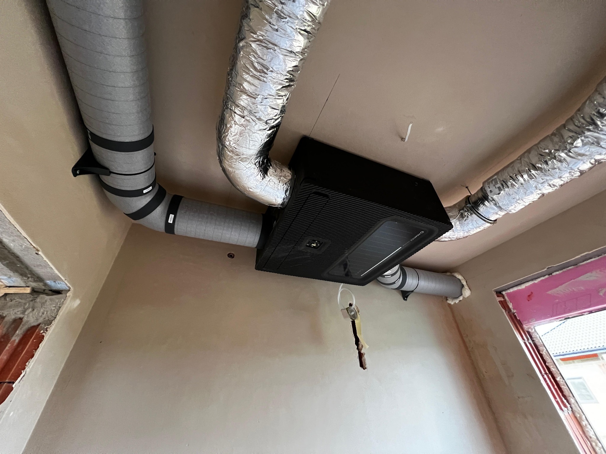 Czarna centrala rekuperacyjna z podłączonymi przewodami wentylacyjnymi w Kobiórze. Instalacja w surowym wnętrzu, widoczne przewody izolowane i aluminiowe.