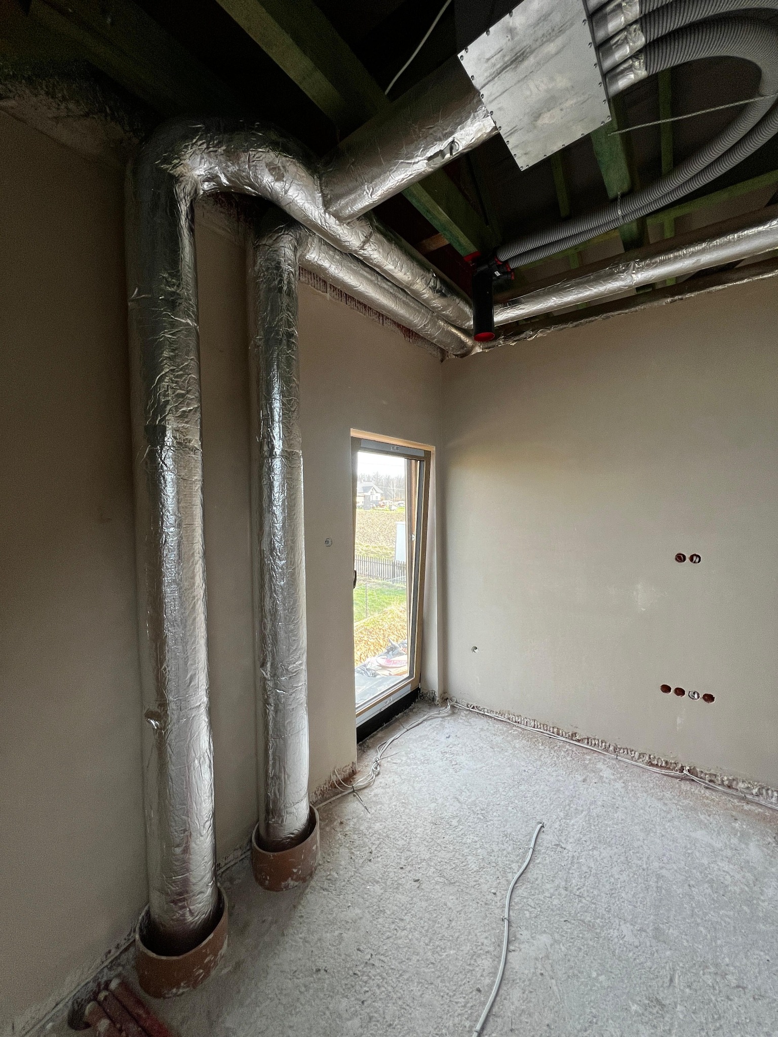 Wnętrze w trakcie montażu instalacji wentylacyjnej z widocznymi izolowanymi przewodami wentylacyjnymi i elementami mocującymi pod sufitem. Okno z widokiem na zewnątrz.