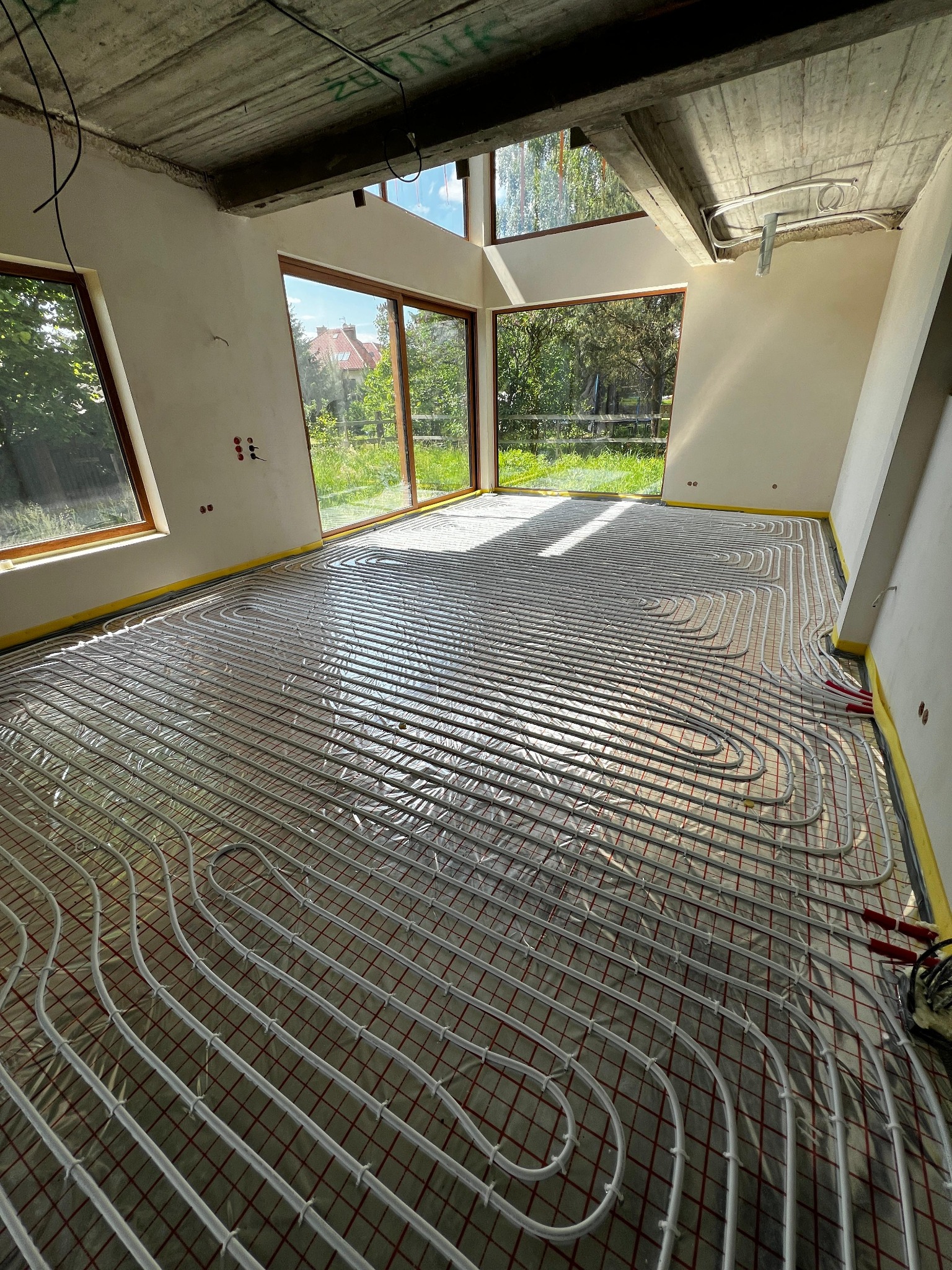 Białe rury ogrzewania podłogowego ułożone na folii w surowym wnętrzu z dużymi oknami i betonowym stropem. Widoczne elementy instalacji przy ścianach.