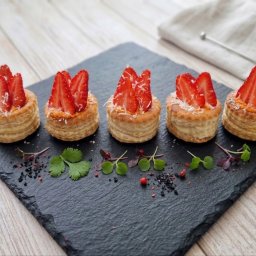 HADAM SPÓŁKA Z OGRANICZONĄ ODPOWIEDZIALNOŚCIĄ - Eleganckie, kruche babeczki z kremem i świeżymi truskawkami, posypane kokosem, ułożone na czarnym, kamiennym talerzu z dekoracją z ziół i przypraw.