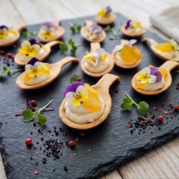 HADAM SPÓŁKA Z OGRANICZONĄ ODPOWIEDZIALNOŚCIĄ - Dekoracyjne finger food na czarnym kamiennym talerzu: mini tartaletki z kremem, owocami i jadalnymi kwiatami, ułożone na drewnianym stole. Elegancki catering.