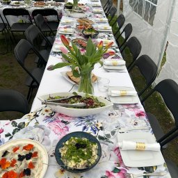 Kuchnia Sztuki - Długi stół pod namiotem, nakryty do przyjęcia z kwiatowym obrusem, zastawą, sałatkami i dekoracjami z kwiatów. Widok z góry na przygotowany event.