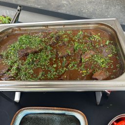 Kuchnia Sztuki - Danie mięsne w ciemnym sosie, posypane zieloną pietruszką, serwowane w metalowym bemarze. Obok miska z sałatą i inne naczynia z jedzeniem.