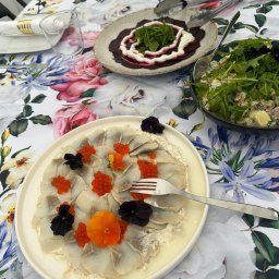 Kuchnia Sztuki - Elegancko podane dania na stole z kwiecistym obrusem: carpaccio z ryby z kawiorem i kwiatami, sałatka z rukolą i buraki z sosem. Catering na przyjęcie.