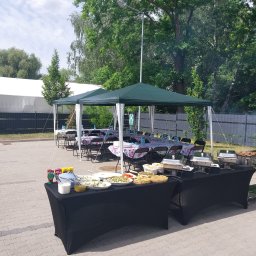 Kuchnia Sztuki - Zewnętrzny catering: stoły pod namiotem z kwiatowymi obrusami, bufet z potrawami, grill. Ustawienie na brukowanym placu, w tle zieleń i ogrodzenie.