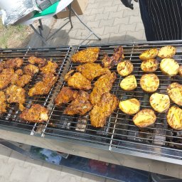 Kuchnia Sztuki - Grill z kurczakiem i ziemniakami w plastrach, przygotowywany na zewnątrz. Widoczny fragment osoby w fartuchu i rękawiczkach. Słoneczny dzień, świeże jedzenie.