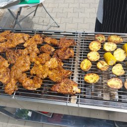 Kuchnia Sztuki - Grill z kurczakiem, ziemniakami i kukurydzą, przygotowywany na zewnątrz. Widoczny fragment osoby w fartuchu. Ujęcie z góry, ostre światło.