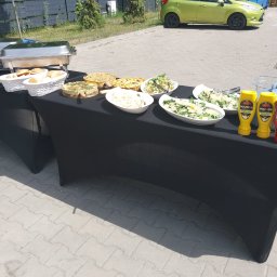 Kuchnia Sztuki - Stół cateringowy z przekąskami: sałatki, tarty, pieczywo, sosy. W tle zielony samochód i fragment osoby w stroju kucharza. Ujęcie z zewnątrz, w słoneczny dzień.