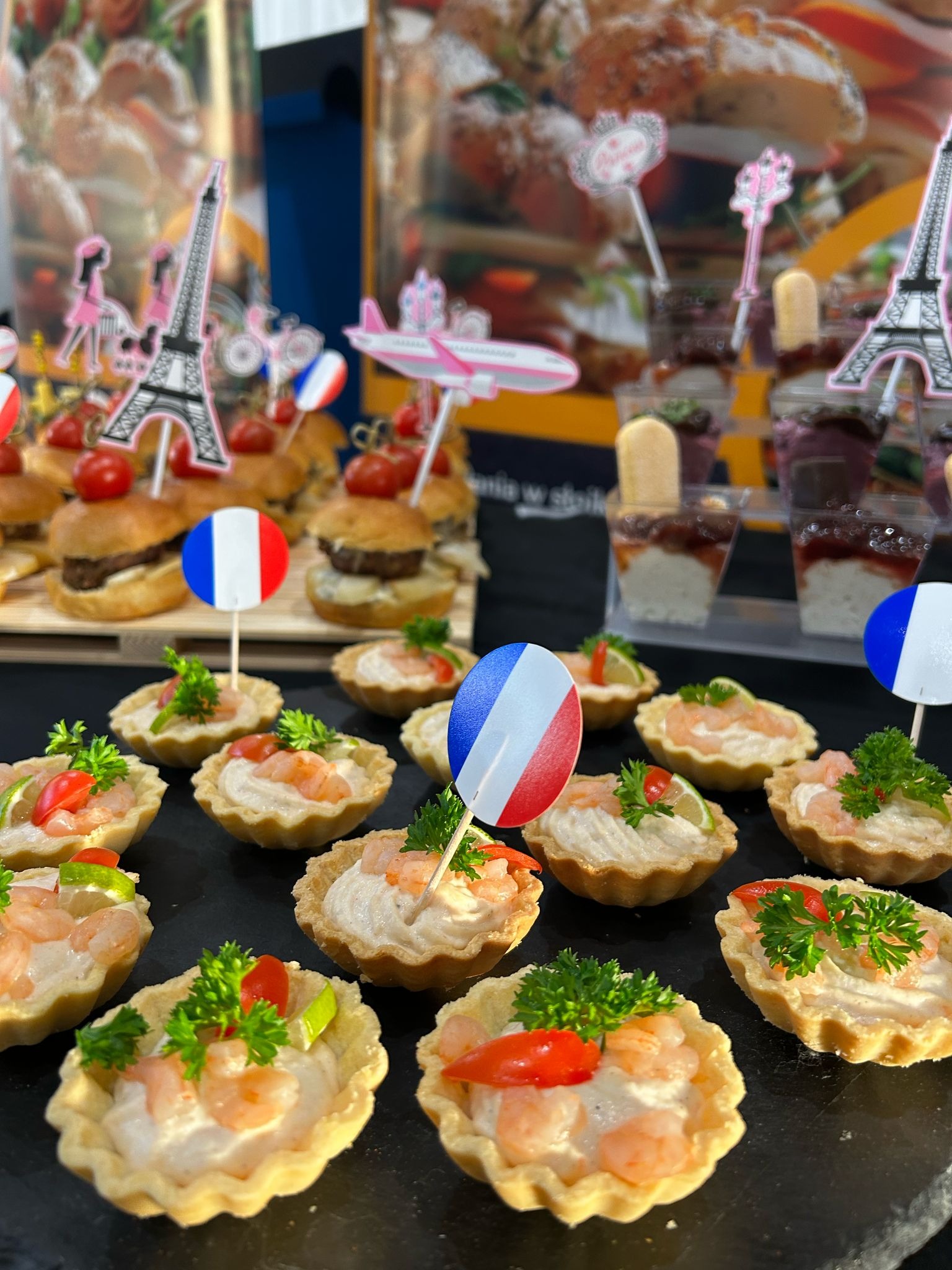 Francuskie przekąski: mini tartaletki z krewetkami, mini burgery i desery w kubeczkach, udekorowane flagami Francji i motywami wieży Eiffla.