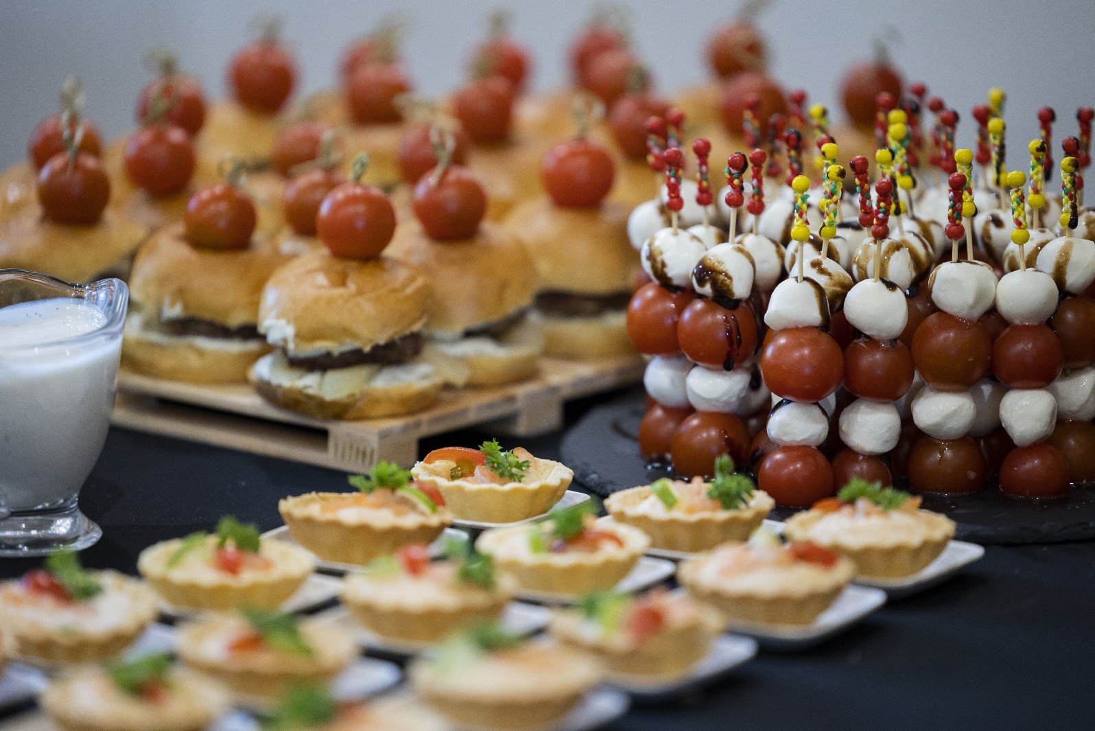 Elegancki catering: mini burgery z pomidorkiem, koreczki z mozzarelli i pomidorów polane sosem balsamicznym oraz wytrawne babeczki na białych talerzykach.