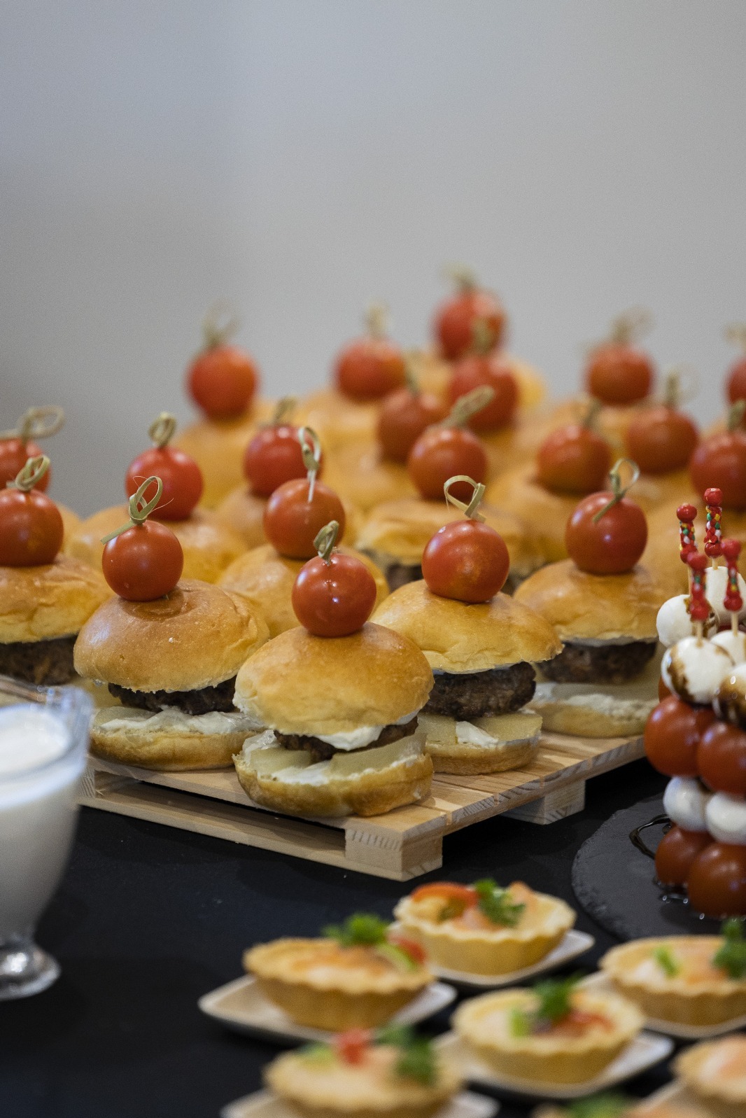 Mini burgery z pomidorkami koktajlowymi na drewnianej palecie, obok tartaletki i szaszłyki caprese. Elegancki catering na przyjęcie.