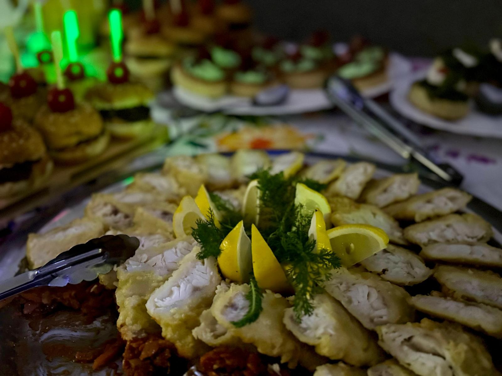 Elegancko podane danie rybne z cytryną i koperkiem na przyjęciu. W tle mini burgery i tartaletki. Srebrne szczypce do nakładania potraw.