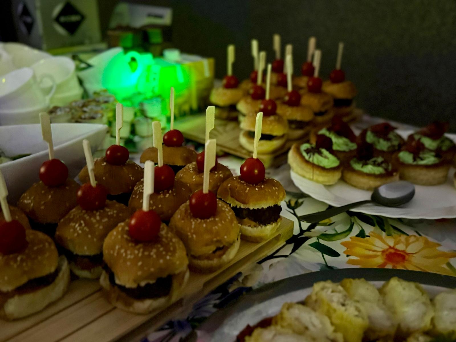 Mini burgery z pomidorkiem koktajlowym i inne przekąski na stole z kwiatowym obrusem, przygotowane na imprezę cateringową. Widoczne oświetlenie dekoracyjne.