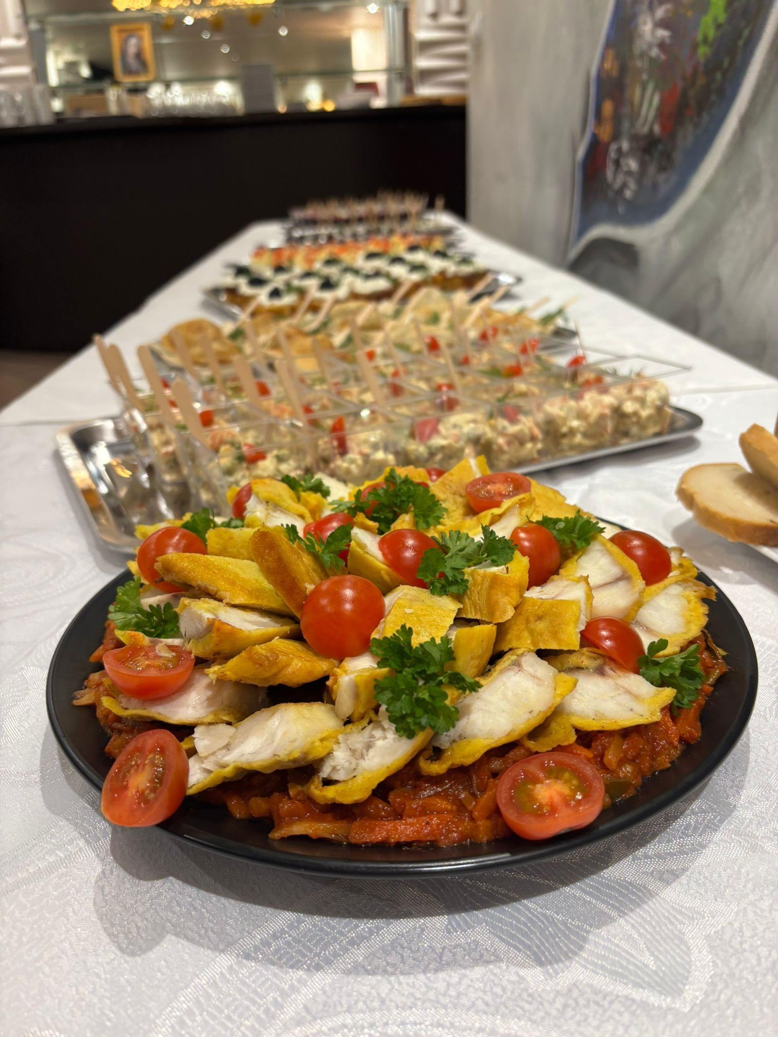 Elegancko podane danie z ryby, pomidorków cherry i zielonej pietruszki na czarnym talerzu. W tle widoczne przekąski na stole z białym obrusem.