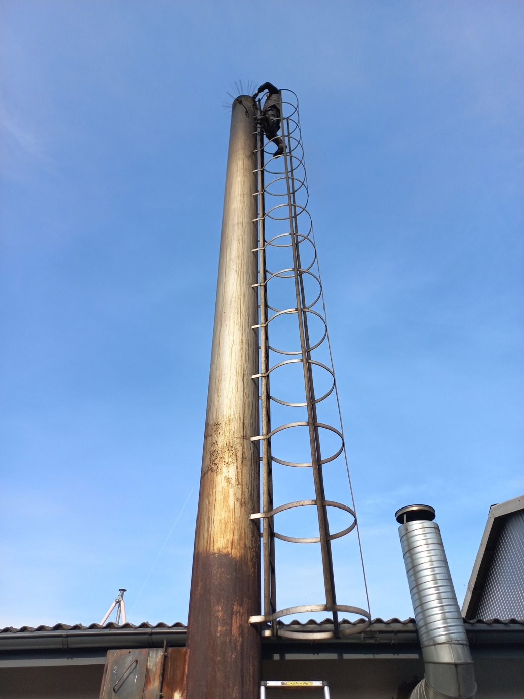 Kominiarz na szczycie wysokiego, metalowego komina z drabinką, na tle błękitnego nieba. Widoczny fragment dachu i drugi komin. Ujęcie z dołu.