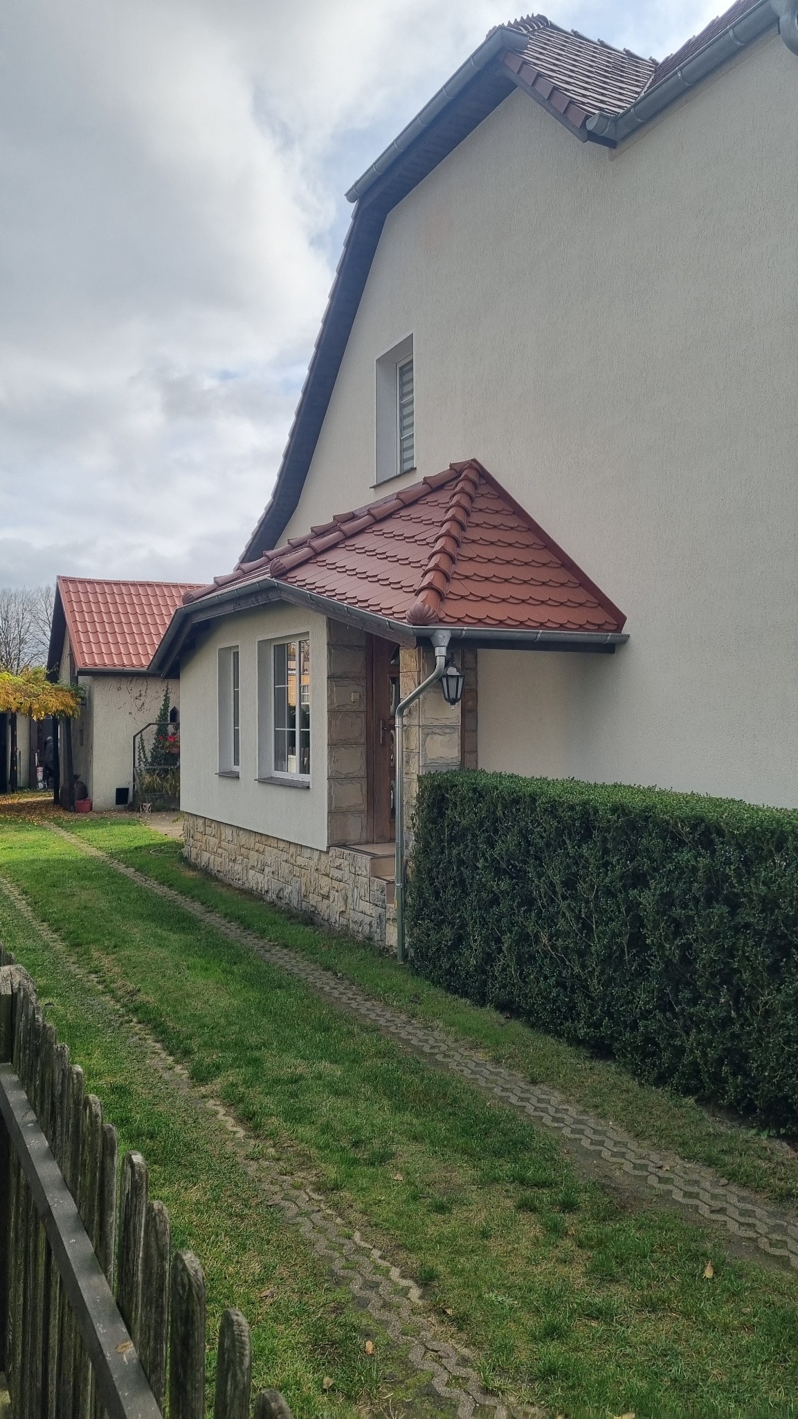 Dom jednorodzinny z czerwonym dachem i rynnami, widok z zewnątrz. Widoczny fragment ogrodzenia i trawnik. Uwagę zwraca oświetlenie zewnętrzne przy wejściu.