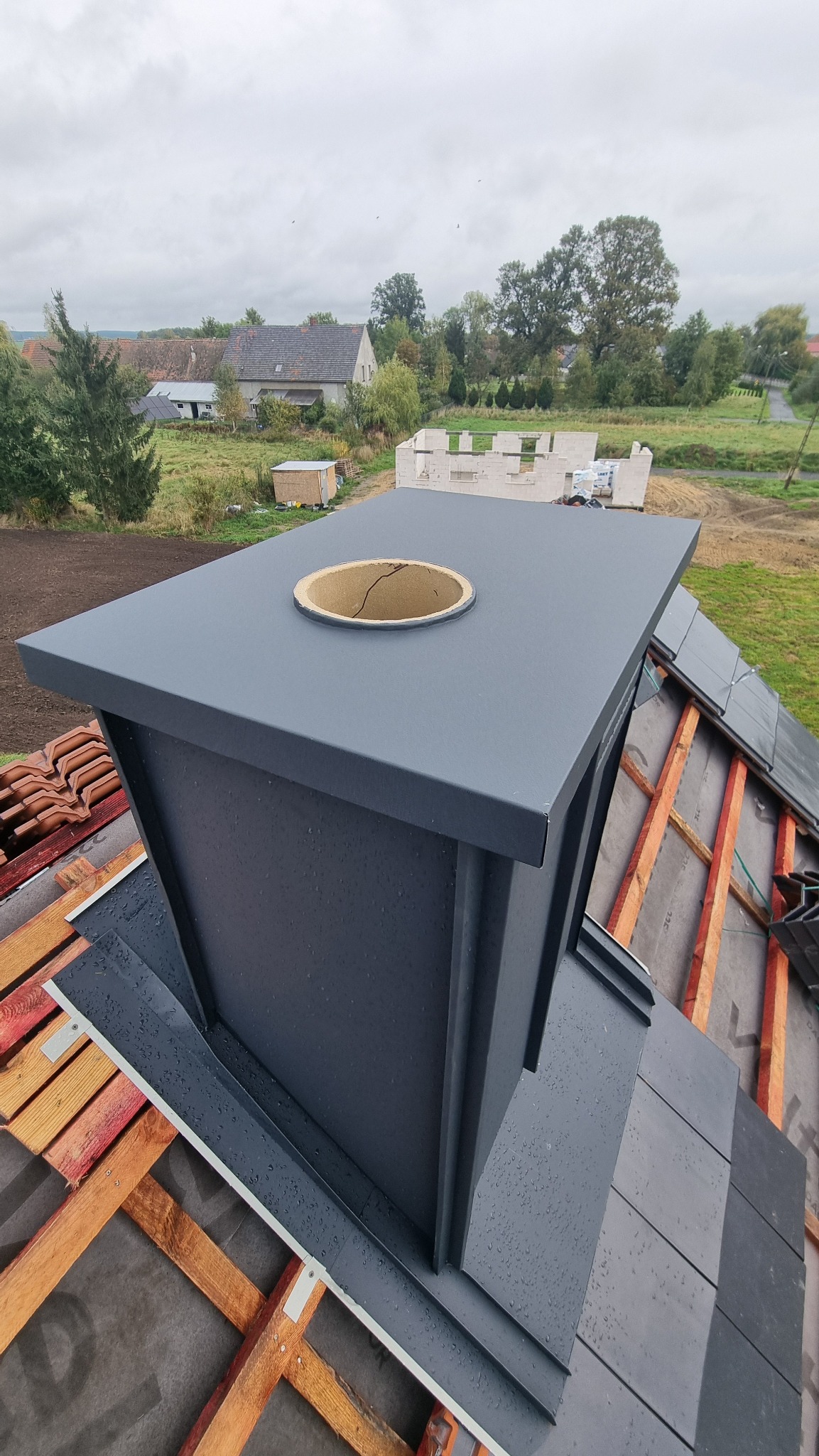 Szary, metalowy komin z otworem na dachu w trakcie budowy, widok z góry. W tle budynki mieszkalne i pochmurne niebo. Widoczne elementy konstrukcji dachu.