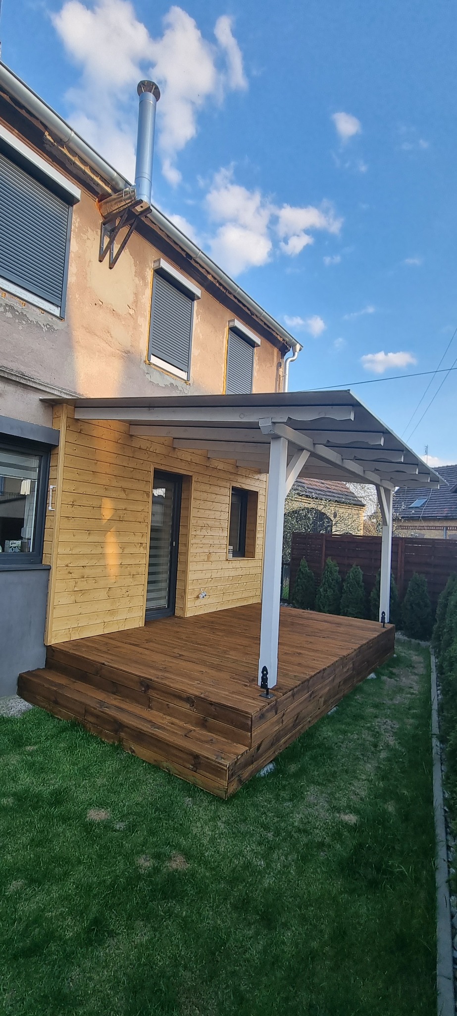 Drewniany taras z zadaszeniem przy budynku z szarymi roletami, widok z poziomu trawnika w słoneczny dzień. Elementy wykończeniowe w kolorze białym.