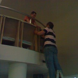 E3 Office Interiors - Dwóch mężczyzn montuje konstrukcję z drewna i metalu na wysokości, jeden na drabinie. Słabe oświetlenie, widok z dołu.