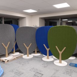 E3 Office Interiors - Kolorowe, tapicerowane panele akustyczne w kształcie drzewek, ustawione w biurze. Widoczny wózek transportowy i paleta, sugerujące montaż lub transport.