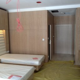 E3 Office Interiors - Jasne drewniane panele ścienne z wbudowanym oświetleniem nad tapicerowanym zagłówkiem łóżka. Podłoga zabezpieczona folią malarską.