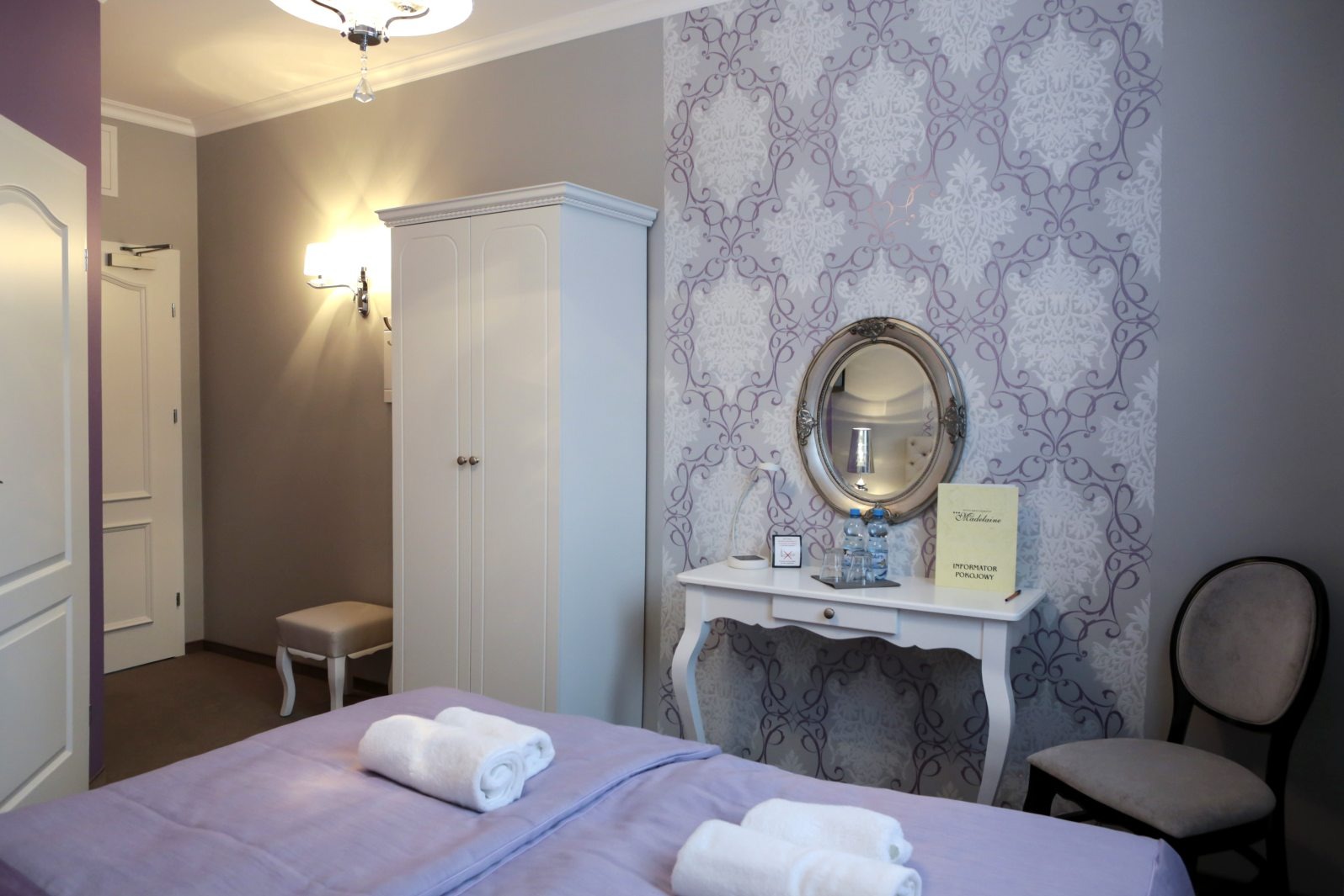 Pokój hotelowy z łóżkiem w liliowym odcieniu, białą szafą i toaletką. Na ścianie wzorzysta tapeta i owalne lustro. Widoczny także krzesło i lampka.