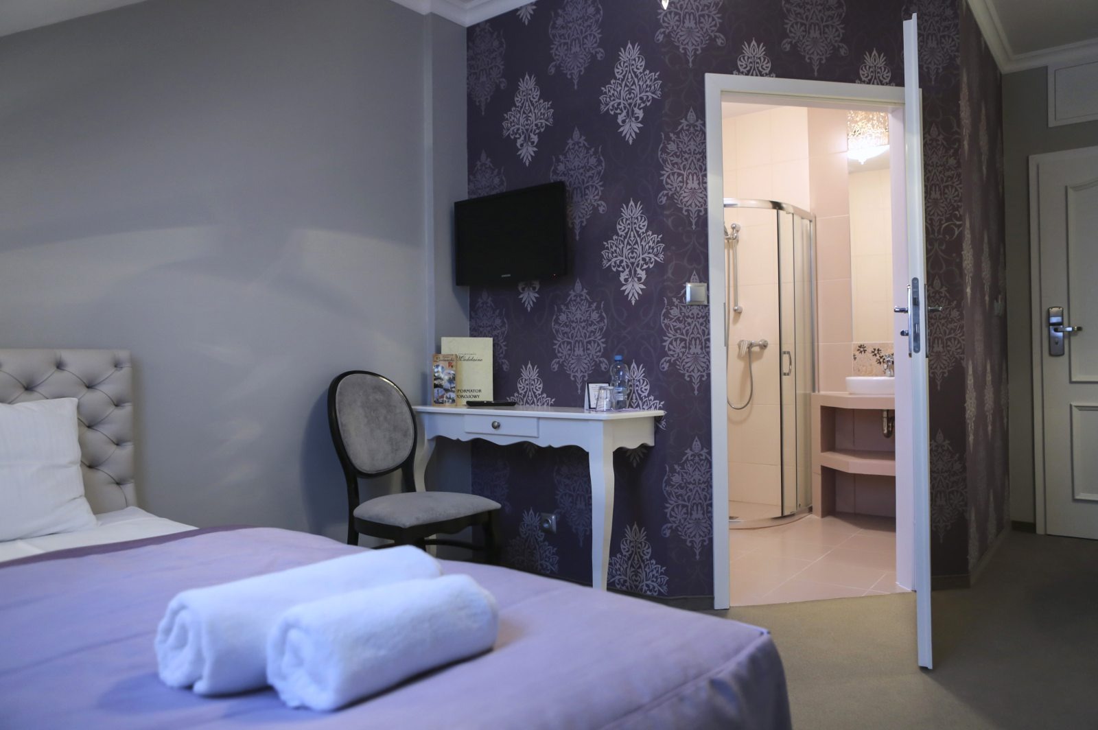 Pokój hotelowy z łazienką w tle: łóżko z fioletową narzutą i ręcznikami, krzesło przy białej toaletce, wzorzysta tapeta i widok na kabinę prysznicową.