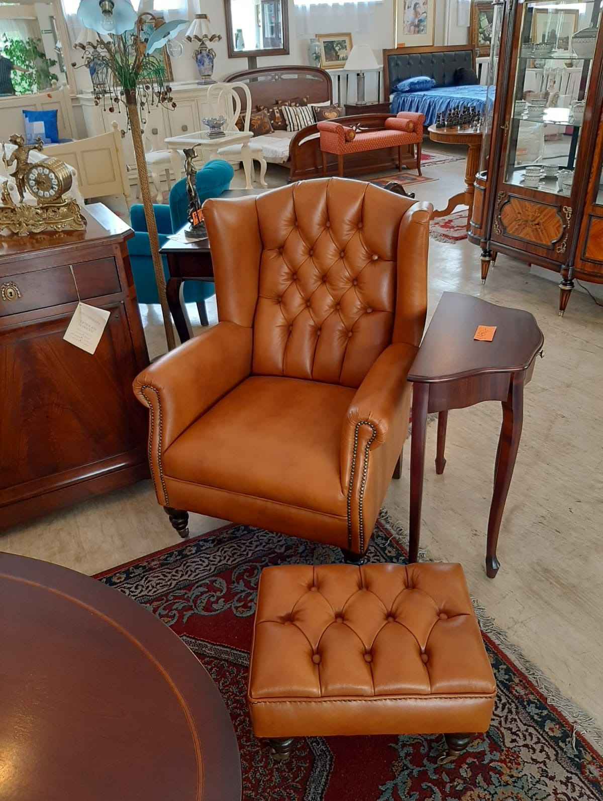 Fotel kapitański w skórze koniakowej z podnóżkiem, pikowany, na tle antycznych mebli i dywanu. Widoczne detale wykończenia i styl.