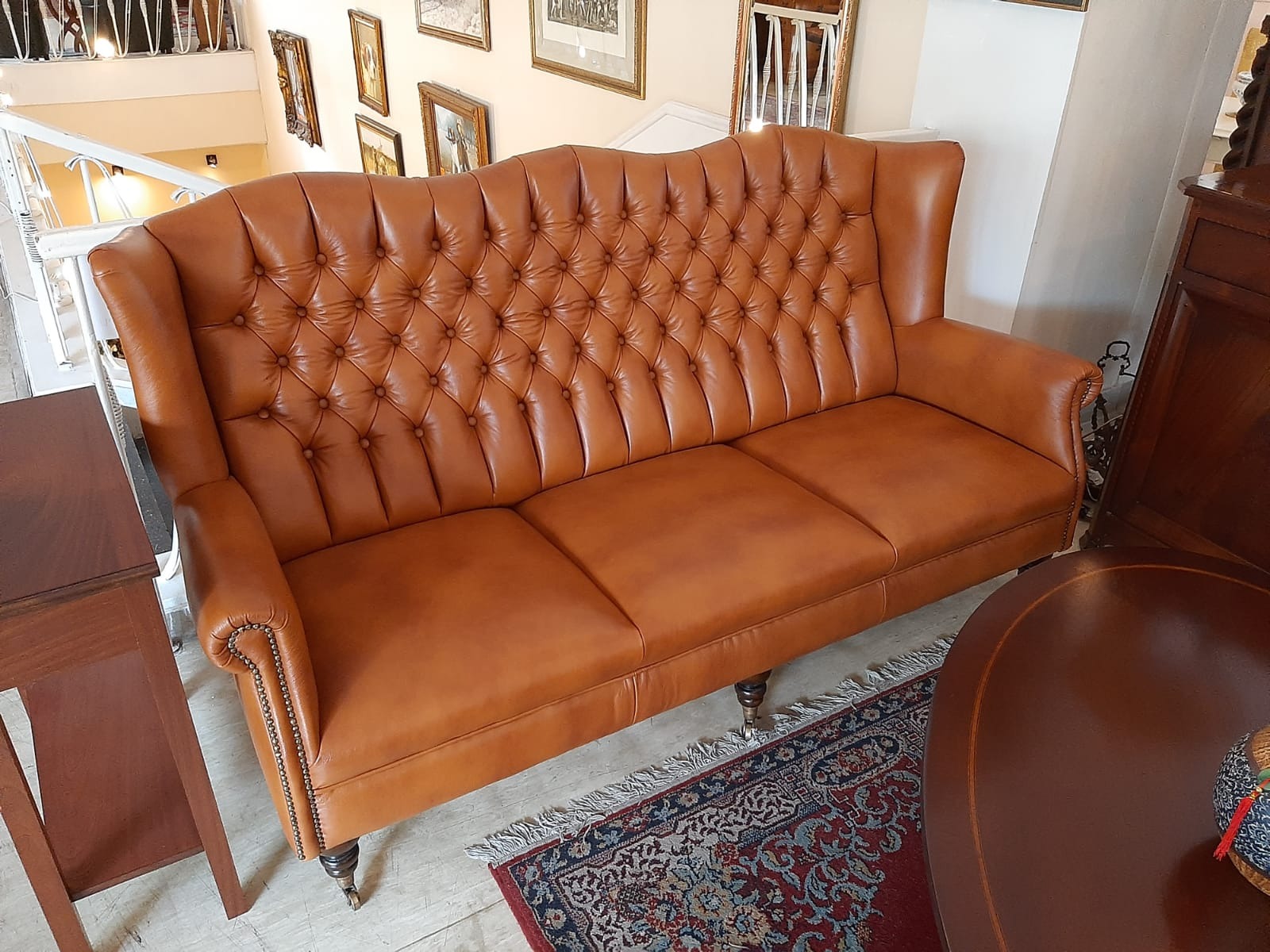 Brązowa, skórzana sofa Chesterfield z pikowanym oparciem, ozdobnymi nitami i toczonymi nogami na tle antycznych mebli i dywanu w salonie.