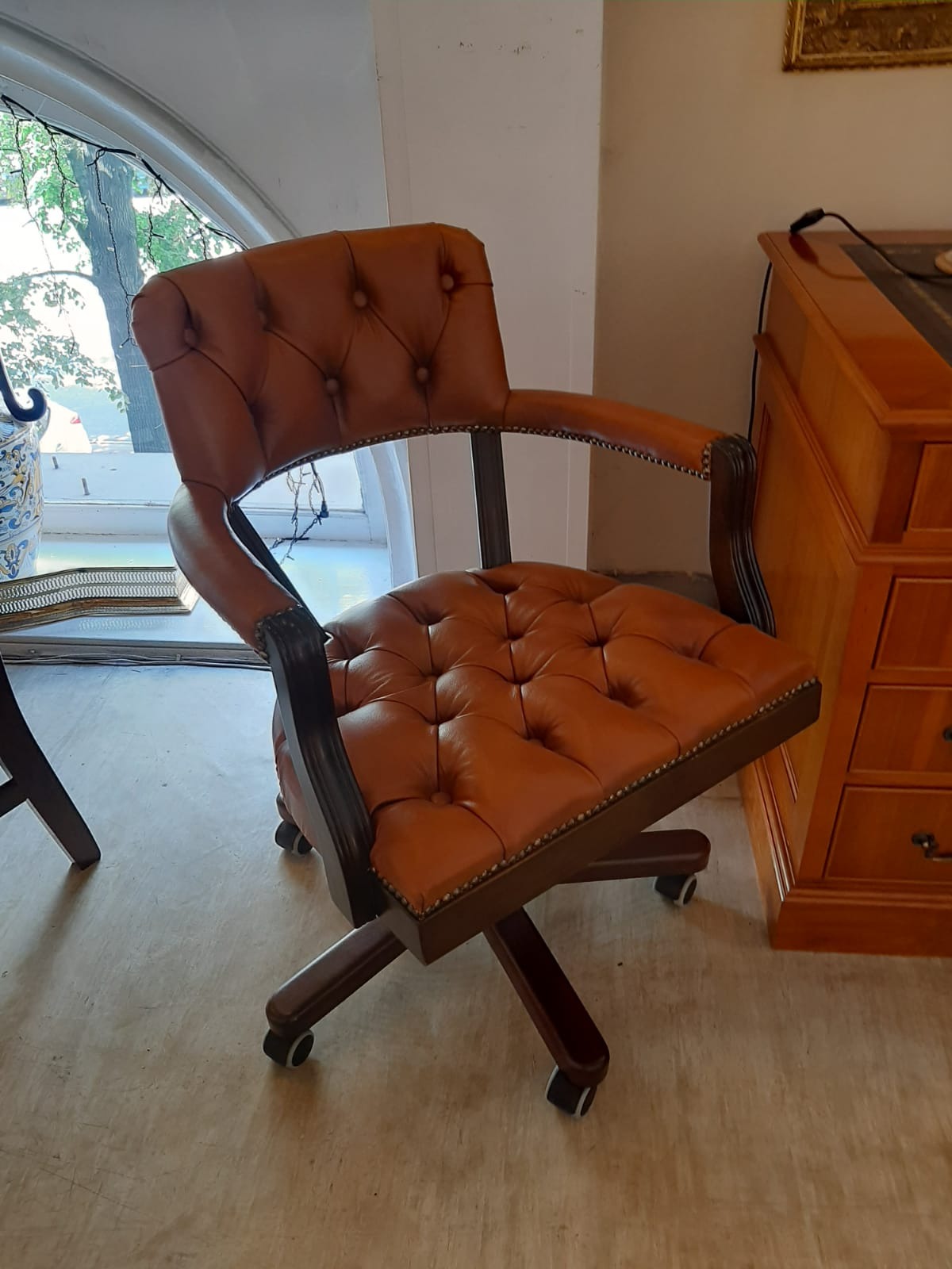 Brązowe, skórzane krzesło biurowe na kółkach, z pikowanym siedziskiem i oparciem, stoi obok drewnianego biurka. Widok z boku, blisko okna.