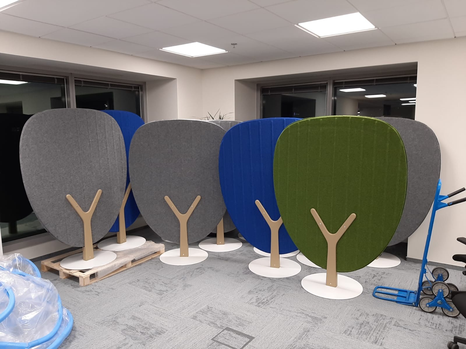 Kolorowe, tapicerowane panele akustyczne w kształcie drzewek, ustawione w biurze. Widoczny wózek transportowy i paleta, sugerujące montaż lub transport.