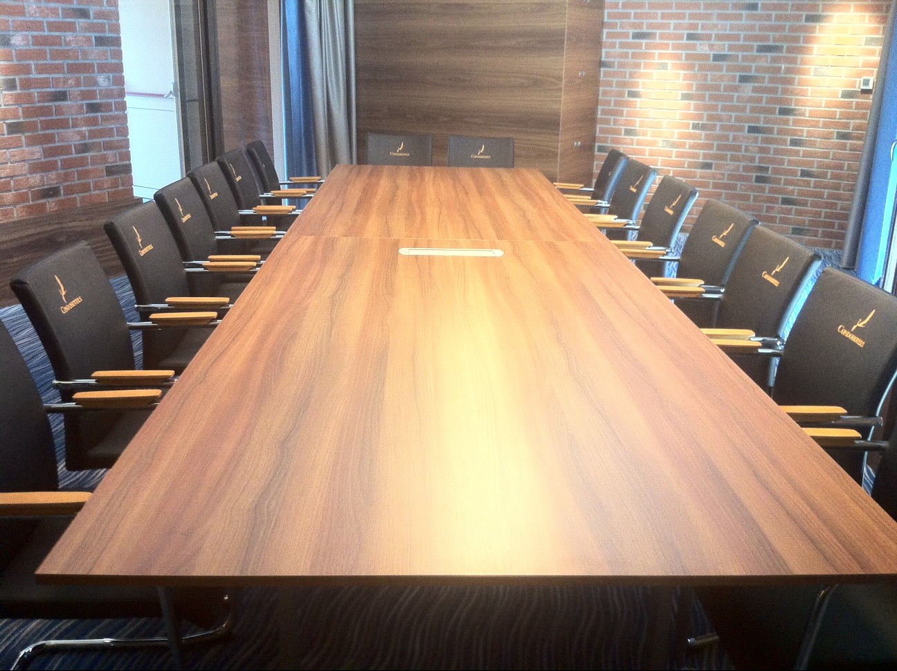 Sala konferencyjna z długim stołem i krzesłami z haftowanym logo. Widok na ceglaną ścianę i zasłony. Oświetlenie górne.