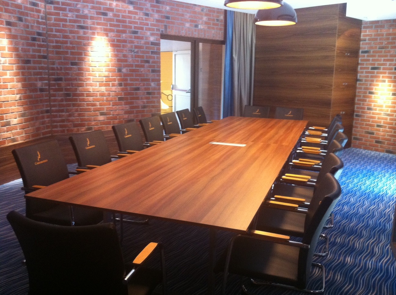 Sala konferencyjna z długim stołem z drewna i krzesłami z logo firmy. Ceglana ściana i nowoczesne oświetlenie tworzą profesjonalną atmosferę.