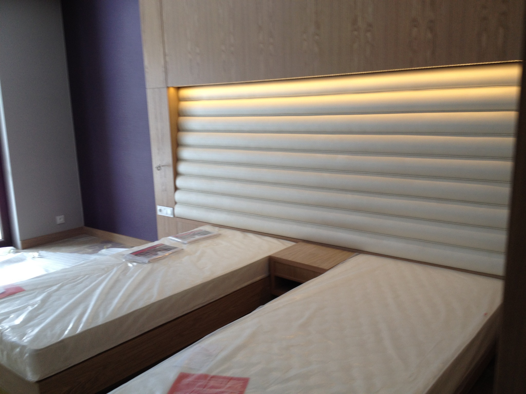 Dwa łóżka hotelowe z tapicerowanymi, podświetlanymi wezgłowiami w odcieniach bieli i beżu, zabezpieczone folią ochronną. Ściana w kolorze fioletowym.