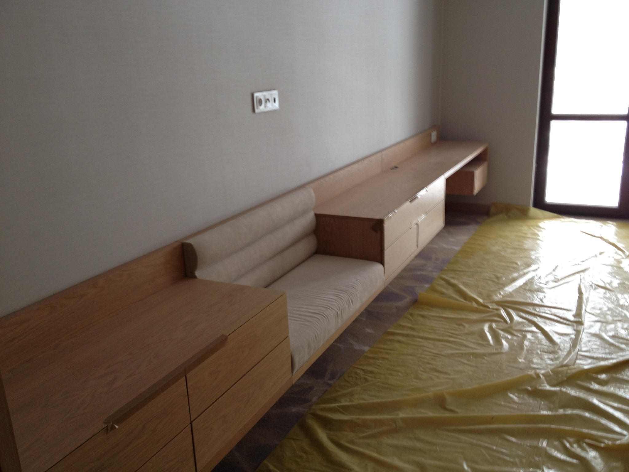 Jasne drewno i tapicerka w zabudowie meblowej z szufladami i siedziskiem, przykryte folią ochronną. Minimalistyczny design, nowoczesne wykończenie.