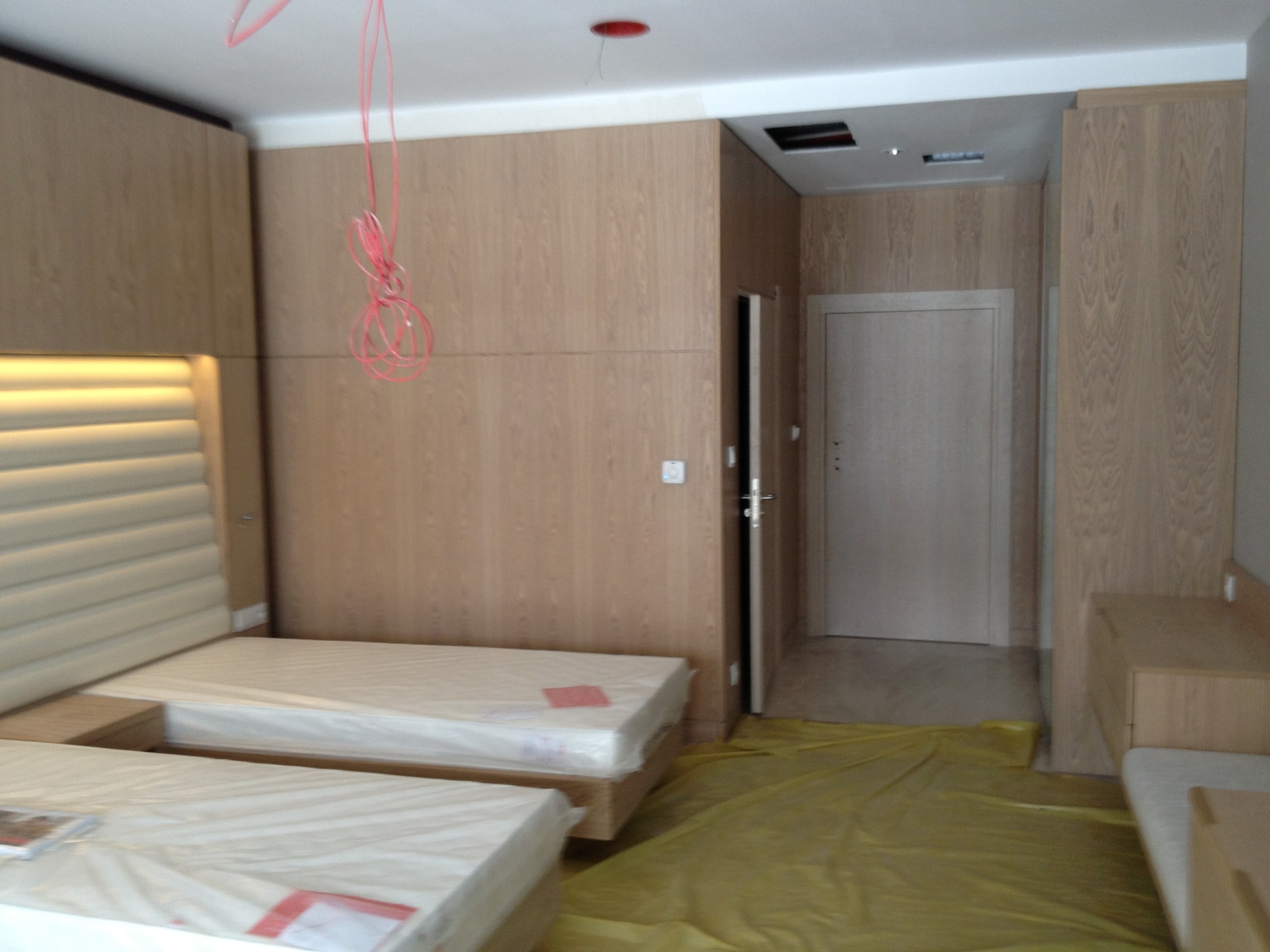 Jasne drewniane panele ścienne z wbudowanym oświetleniem nad tapicerowanym zagłówkiem łóżka. Podłoga zabezpieczona folią malarską.