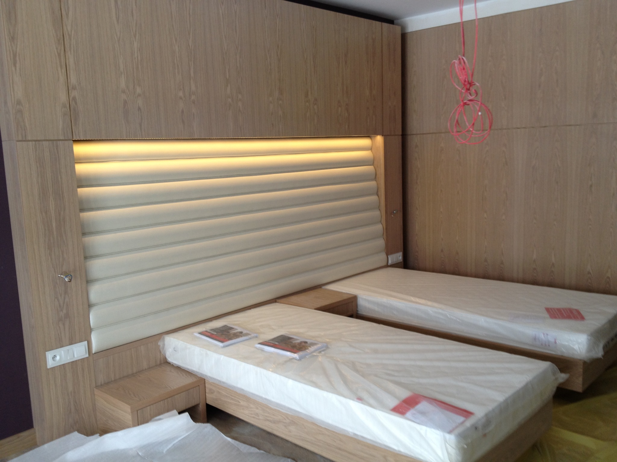 Dwa łóżka z tapicerowanym zagłówkiem w jasnej kolorystyce, z drewnianą zabudową i oświetleniem LED. Na materacach leżą książki, a całość jest jeszcze w folii ochronnej.